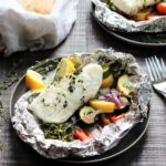 Opened foil packets with grilled halibut on top of vegetables with bread basket in the background on a wooden surface with forks on the side.
