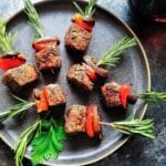 Bitesize filet mignon appetizer skewers with rosemary served on a dark gray plate with red wine.