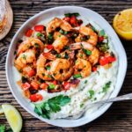 Plump cajun shrimp and grits garnished with parsley and lemon wedges served in bowl on wooden board.