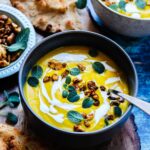 Vibrant turmeric cauliflower soup topped with sage, toasted cashews and coconut cream in a dark gray bowl on a rustic wooden board.