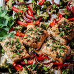 Four salmon fillets topped with sliced green onions in the middle of charred, roasted veggies on a sheet pan.