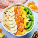 Fruit topped smoothie in a white bowl with granola, mango, orange slices and orange printed napkin on the side.