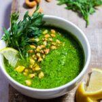 Close up of carrot top pesto garnished with chopped cashews, carrot greens and lemon on a wooden board.
