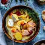 Spoon in brown bowl full of fish chowder garnished with thyme and fennel fronds with a blue napkin on the left side.