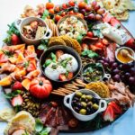 Fully assembled round charcuterie and cheese board with wine and naan on the side on a white surface.