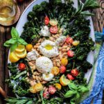 Assembled charred broccolini salad topped with beans, cherry tomatoes, burrata, parmesan and fresh herbs on a large white platter.