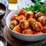 Meatballs served in a shallow bowl with extra buffalo sauce, celery sticks and a glass of beer in the background.