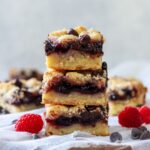 Stack of 3 chocolate raspberry bars on a cheesecloth lined surface with chocolate chips and fresh raspberries on the sides.