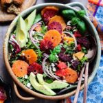 Cara cara and blood orange slices arranged over baby greens with avocado, goat cheese and dried cranberries in a large bowl.
