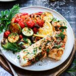 Oven baked halibut and vegetables served on a white plate with the fork to the side on a wooden charger.