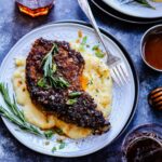 Pretzel crusted pork chop and polenta served on a white plate with glasses and a fork to the side on a dark blue textured surface.