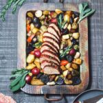 Juicy pork tenderloin sliced and served with potatoes, carrots and brussels sprouts on a metal platter on a wooden surface.