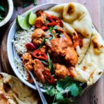 Butter chicken and cauliflower served with naan in a bowl over rice garnished with cilantro, green onion and fresno peppers on a wooden surface.