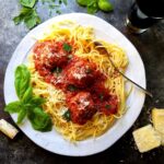Spaghetti, meatballs and tomato sauce on a round white plate with fresh herbs on a dark metallic surface with red wine and parmesan cheese on the side.